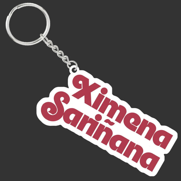 Maqueta digital en alta definición sobre fondo oscuro que detalla el llavero de Ximena Sariñana con su argolla metálica y el dije acrílico troquelado con letras rojas.