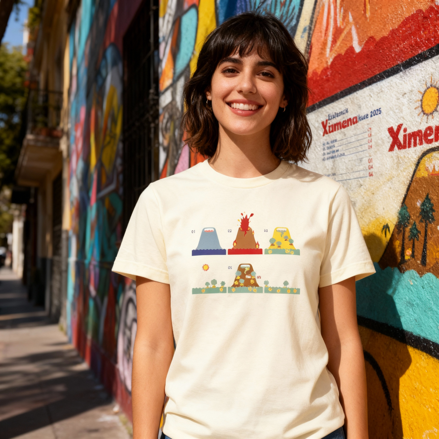 Fotografía en plano medio (medium shot) de una modelo mujer sonriendo de frente, vistiendo la playera color crema de Ximena Sariñana con ilustraciones frontales ante una pared con arte urbano.