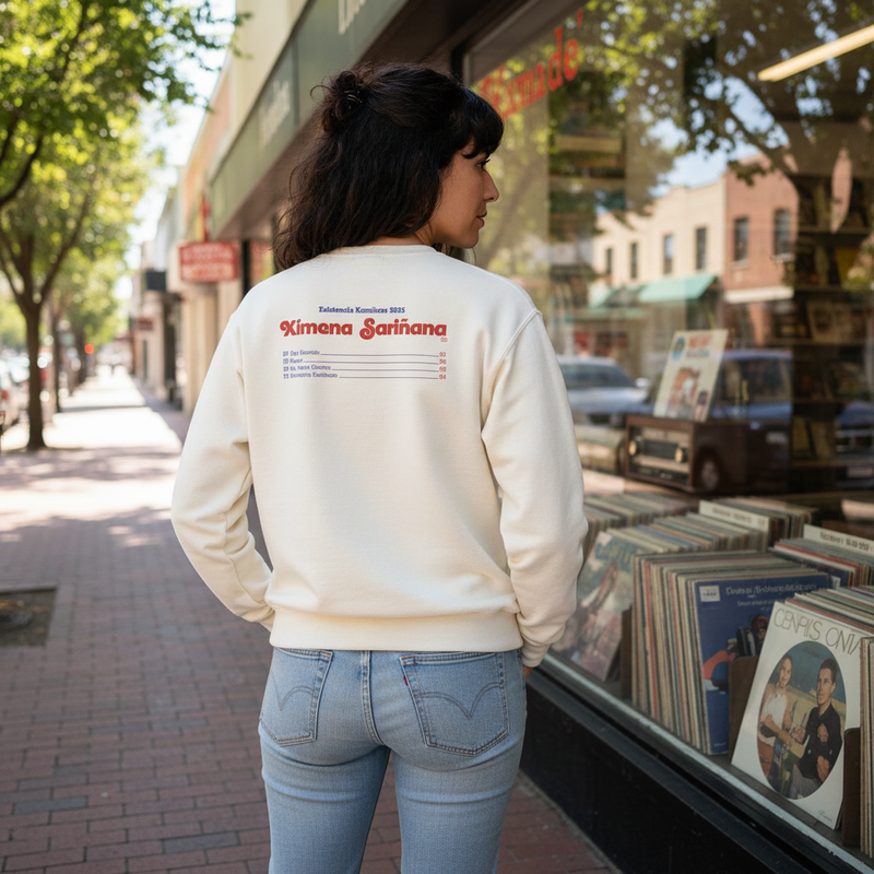 Fotografía en plano medio (medium shot) desde atrás de una modelo caminando por la acera de una calle, exhibiendo el diseño posterior de la sudadera oficial de Ximena Sariñana.