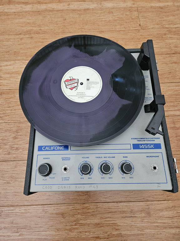Fotografía que muestra uno de los discos de vinilo color morado y negro de Miguel Bosé colocado sobre el plato de un tocadiscos portátil de estilo clásico.