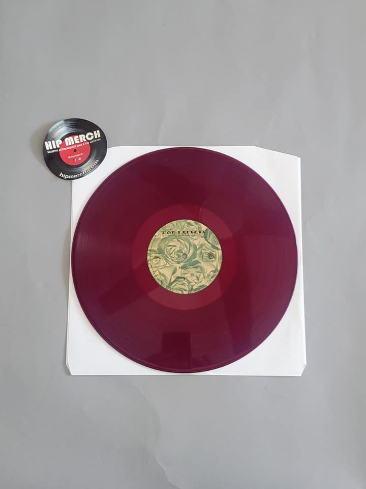 Fotografía completa que muestra el disco de vinilo color magenta de Mon Laferte Vol. 1 reposando sobre su funda protectora de papel blanco junto al pin decorativo.