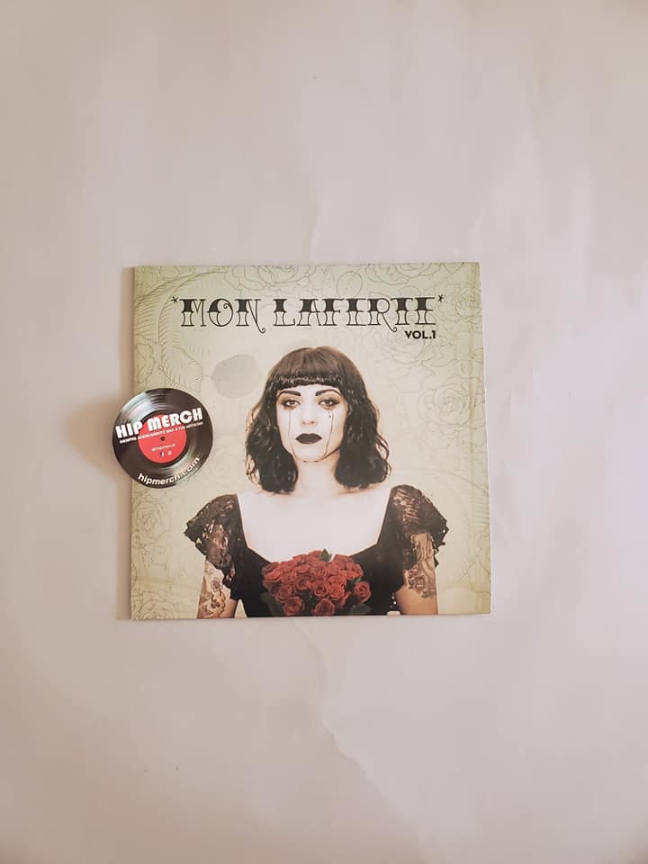 Fotografía en primer plano de la portada frontal del disco de vinilo Mon Laferte Vol. 1 con un pin decorativo circular colocado sobre el lado izquierdo.