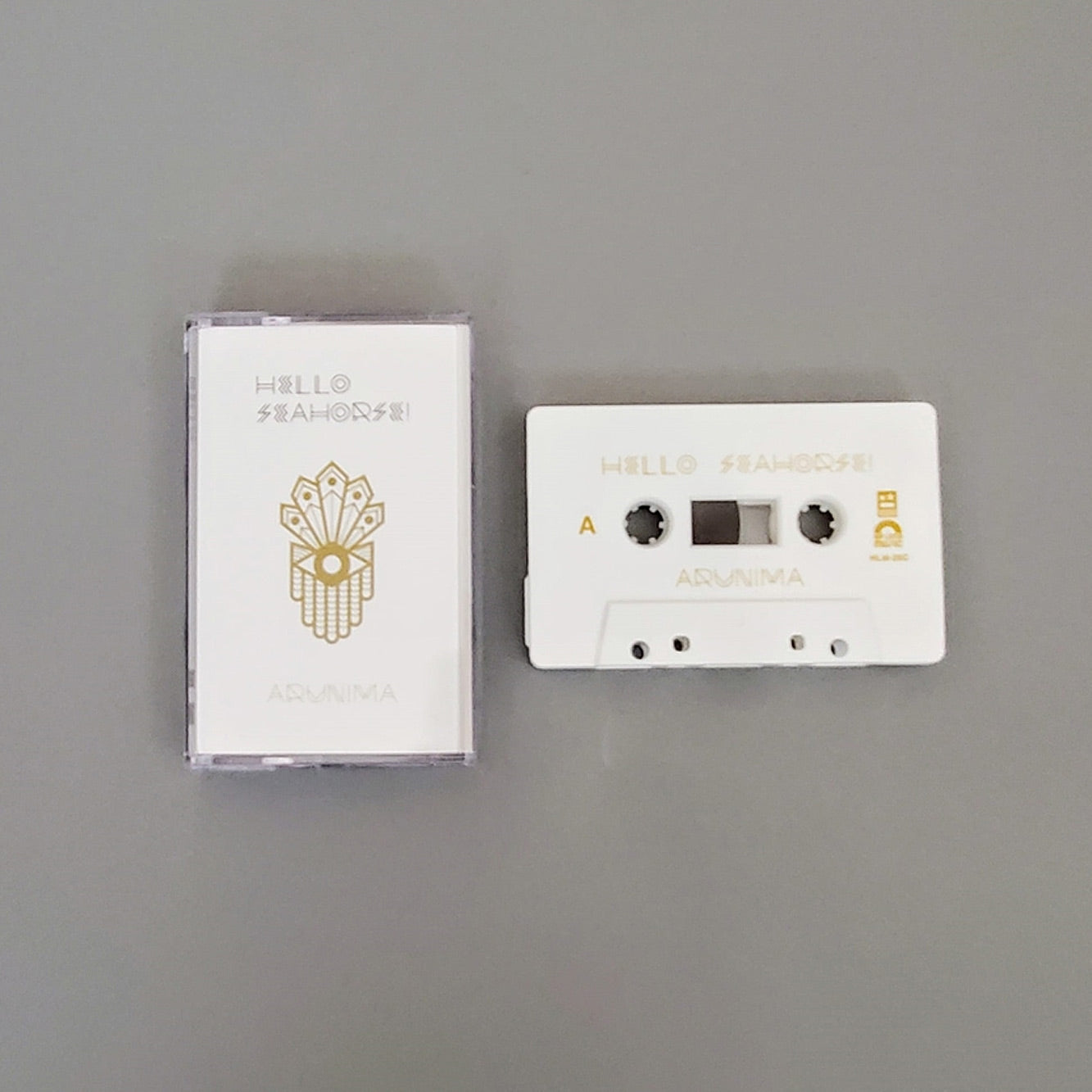 Fotografía de la edición limitada en cassette del álbum Arunima de Hello Seahorse!, exhibiendo la cinta física en color blanco con detalles dorados junto a su estuche oficial.