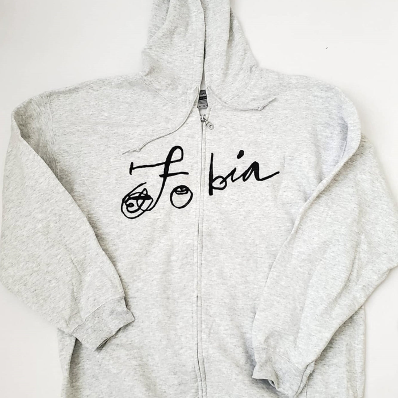 Fotografía de la vista frontal de la sudadera gris claro oficial de Fobia, mostrando el diseño con cierre, capucha y el logotipo de la banda estampado en color negro.